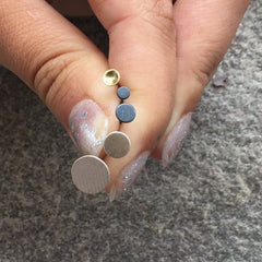 A close-up of a hand with a stack of small round stud earrings in gold, black, and silver tones against a textured gray background.